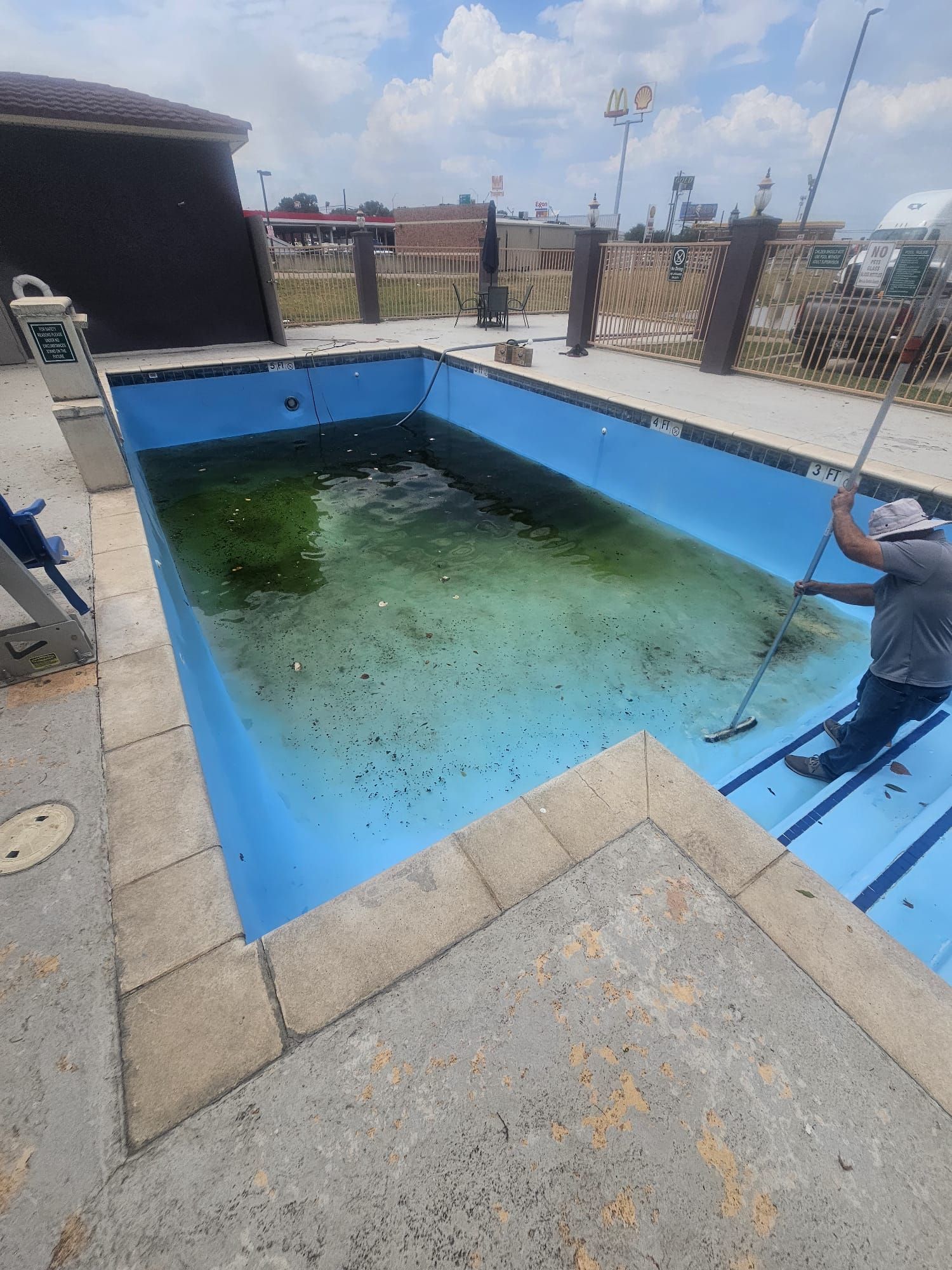 Man cleaning a neglected, algae-filled blue pool with a long-handled brush on a sunny day.