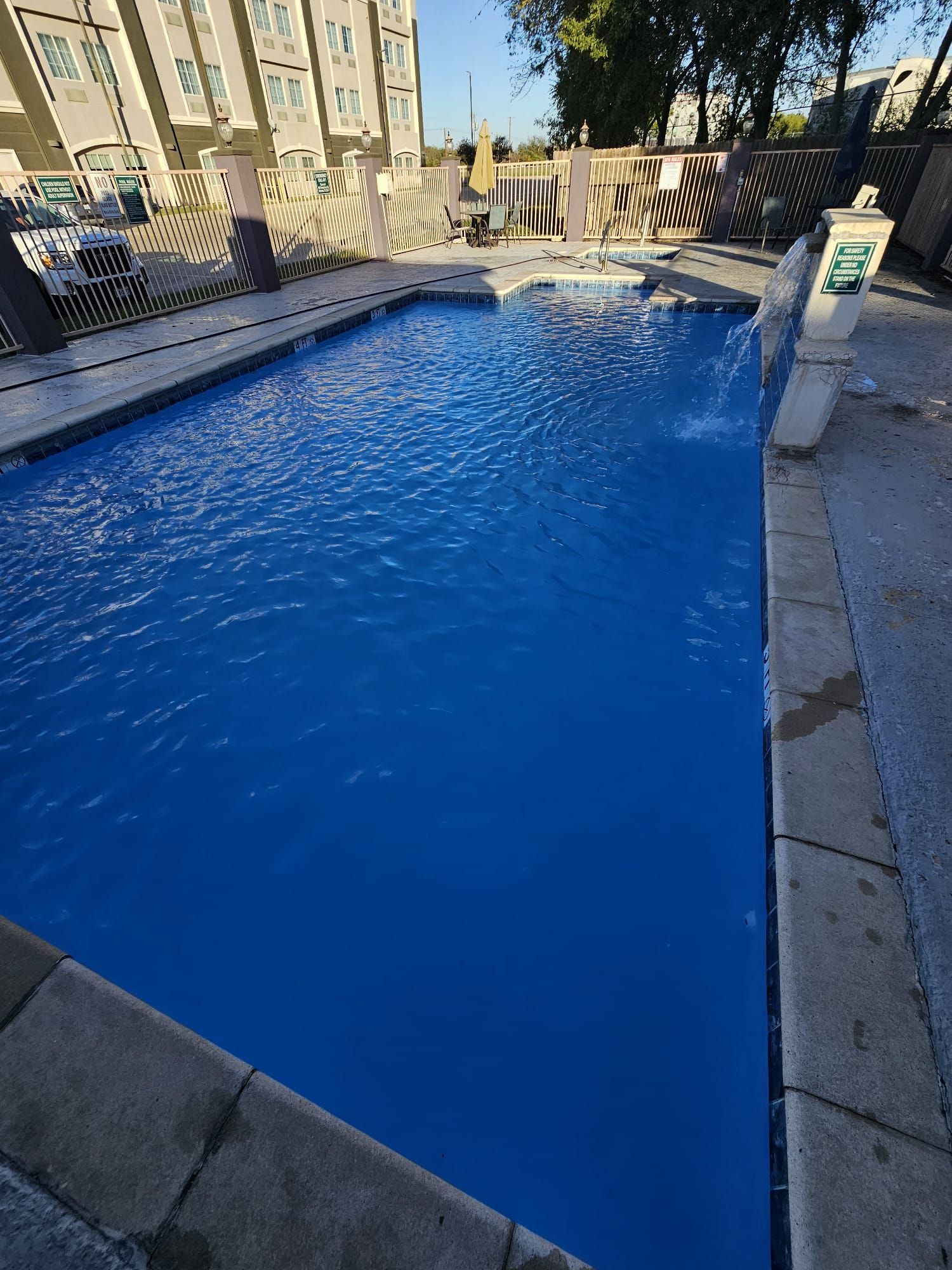Rectangular outdoor swimming pool with bright blue water, surrounded by concrete and a fence.