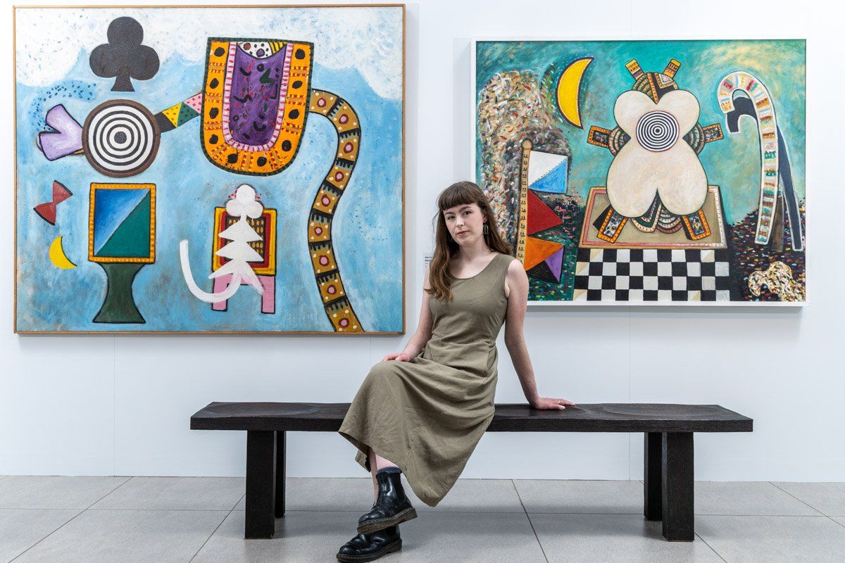 Photo of woman sitting on a bench with two colourful paintings on the white wall behind