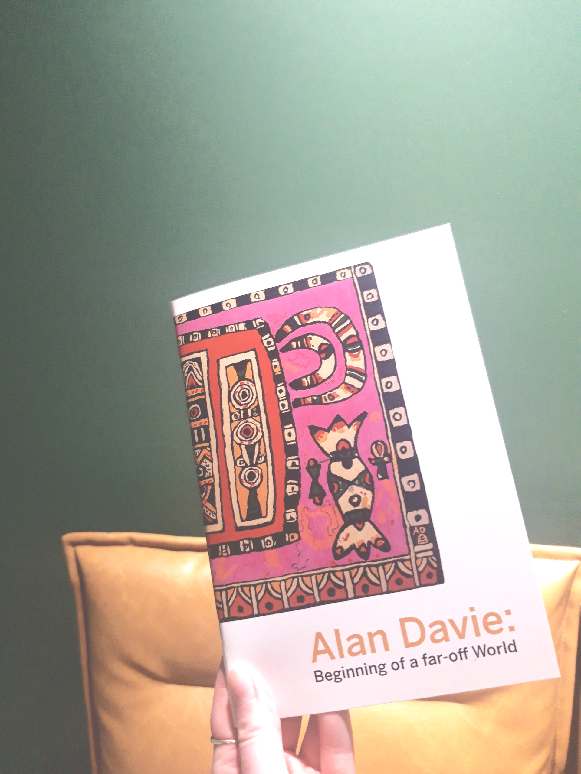 Hand holding book with pink photograph and text 'Alan Davie' in front of green BBC wall