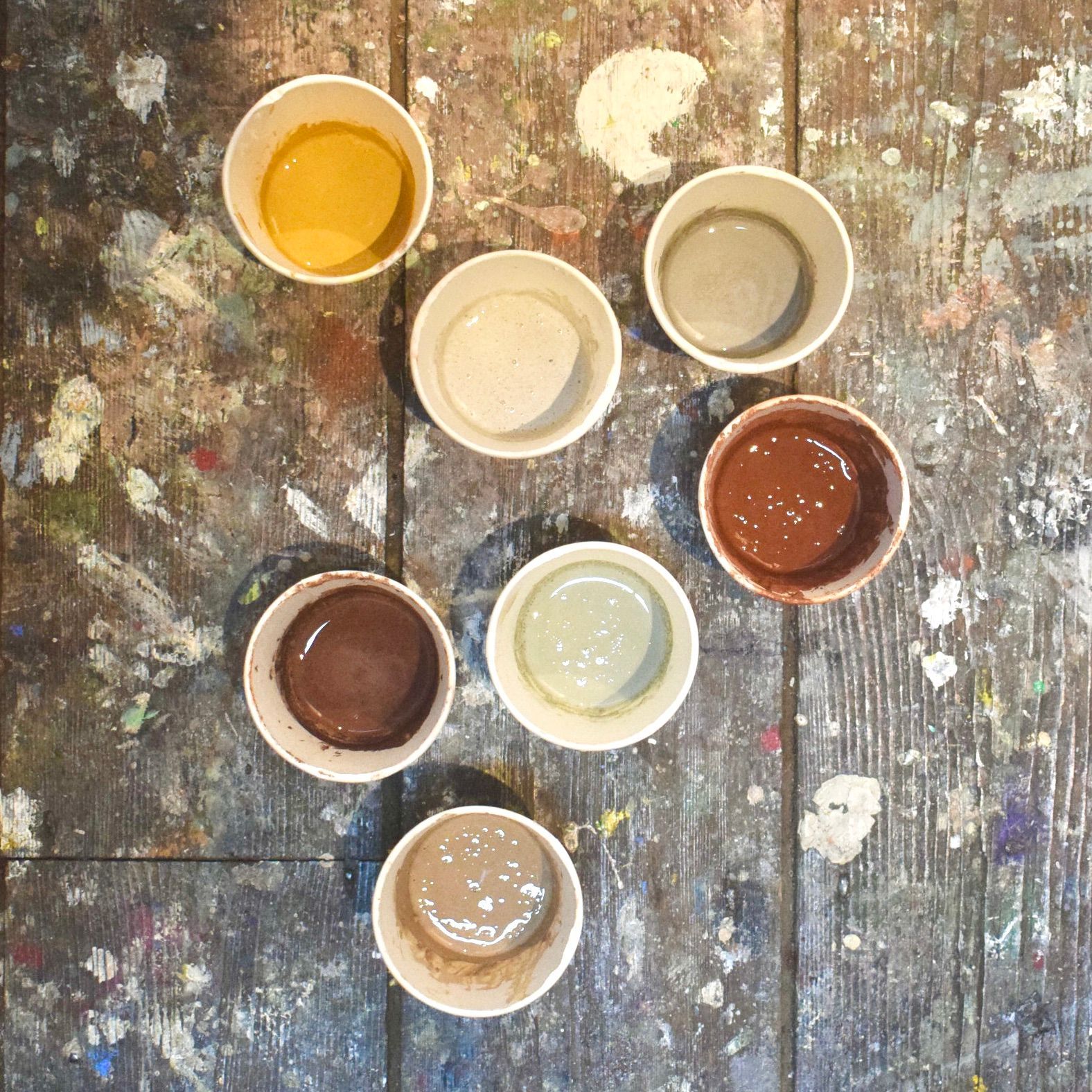 Photograph of 7 cups of coloured liquid paint on a wooden floor