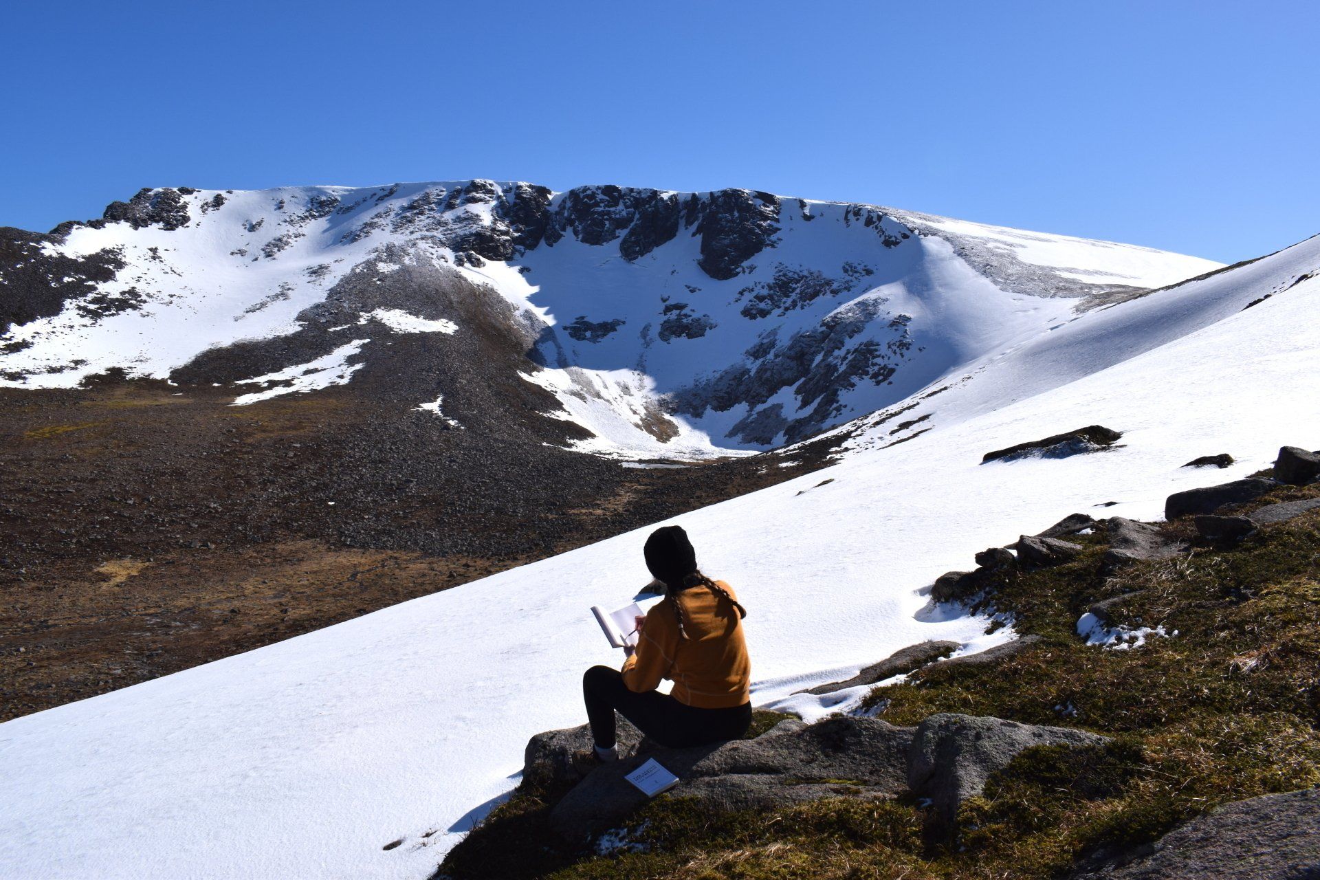 Photo of woman sitting on rock in front of mountain covered in white snow, with blue sky