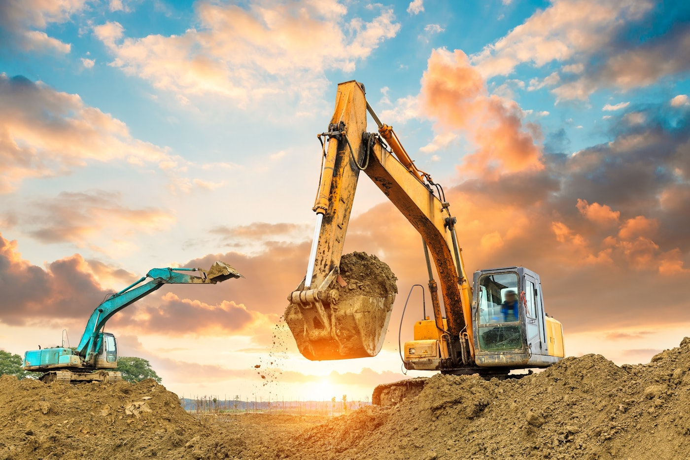 Two excavators are working on a construction site at sunset.