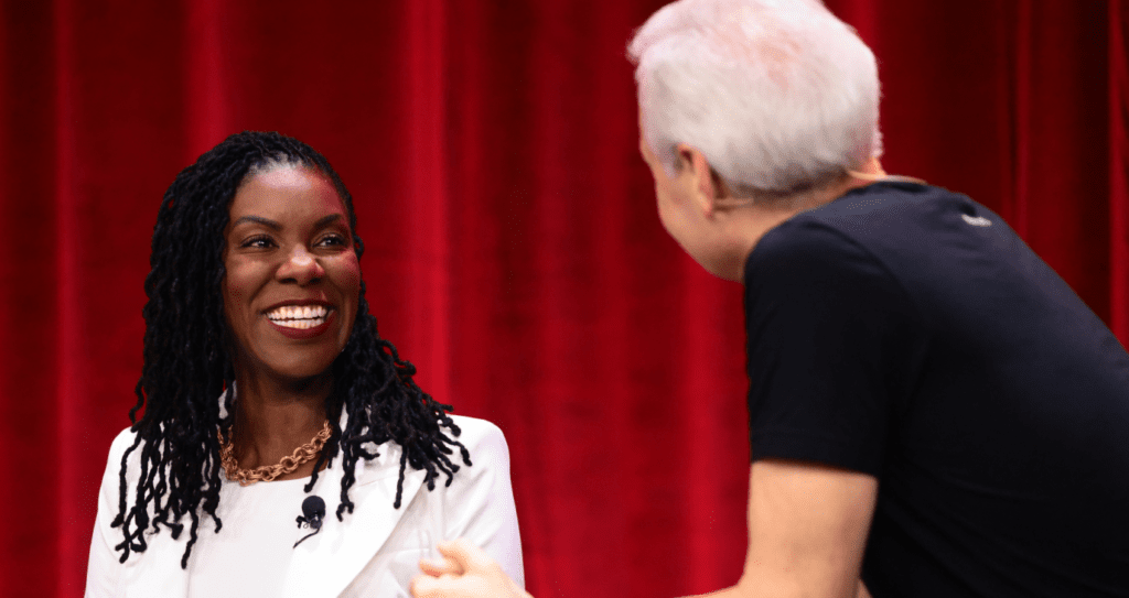 Two women are talking to each other on a stage in front of a red curtain.