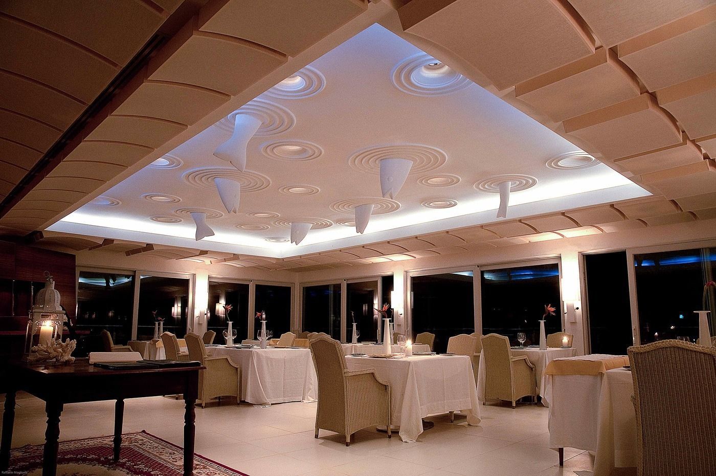 Interno del ristorante con tavoli apparecchiati per la cena, soffitto in legno e opere d'arte alle pareti.