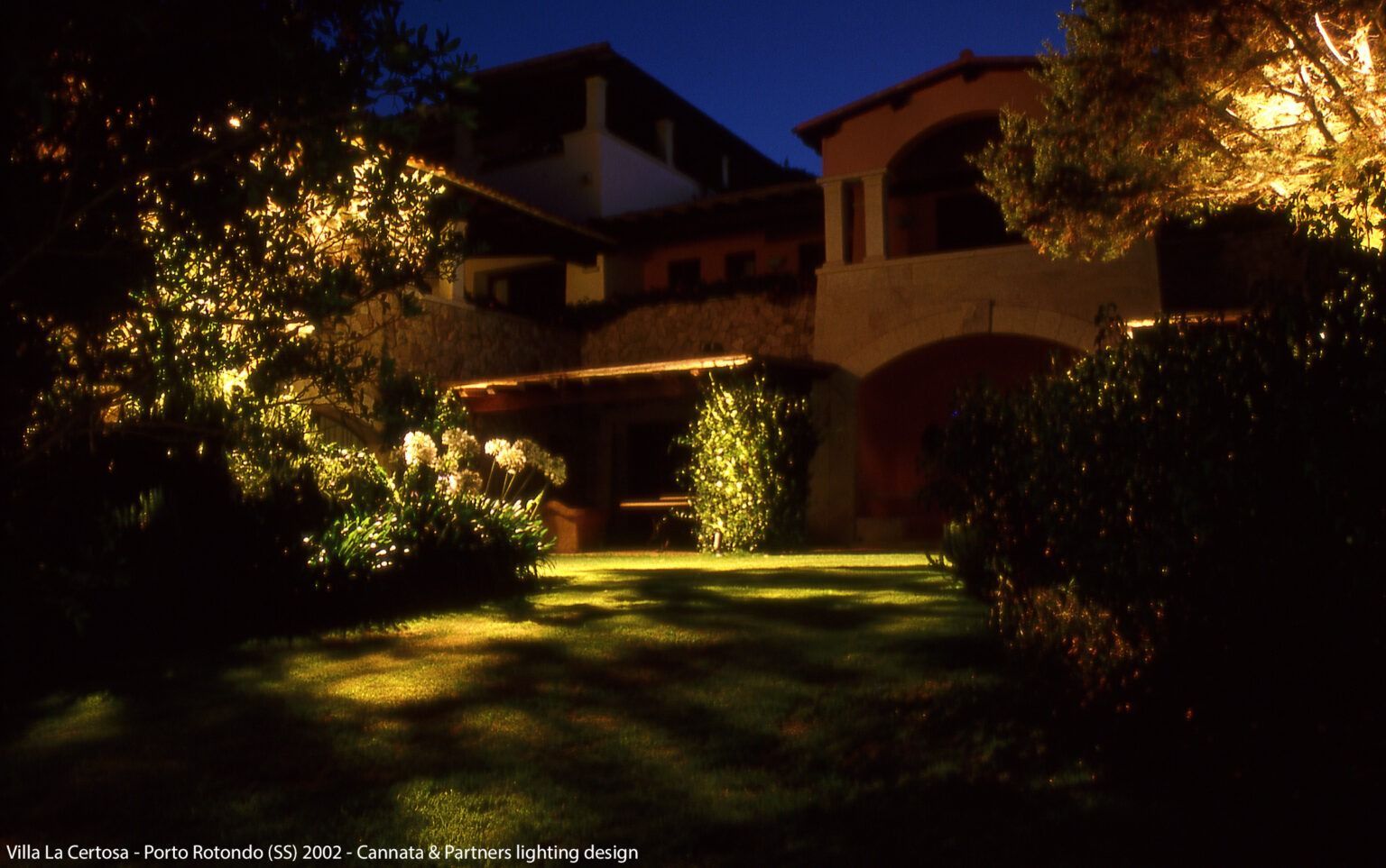 Vista notturna di una casa con illuminazione esterna, che illumina un prato e il fogliame.
