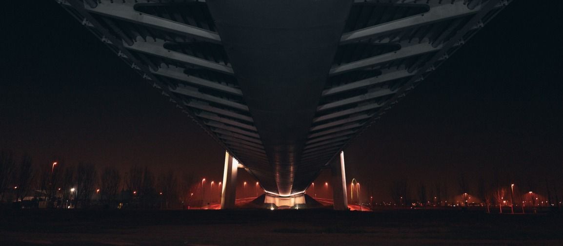 Vista notturna da sotto un ponte; luci sulla riva, acqua scura.