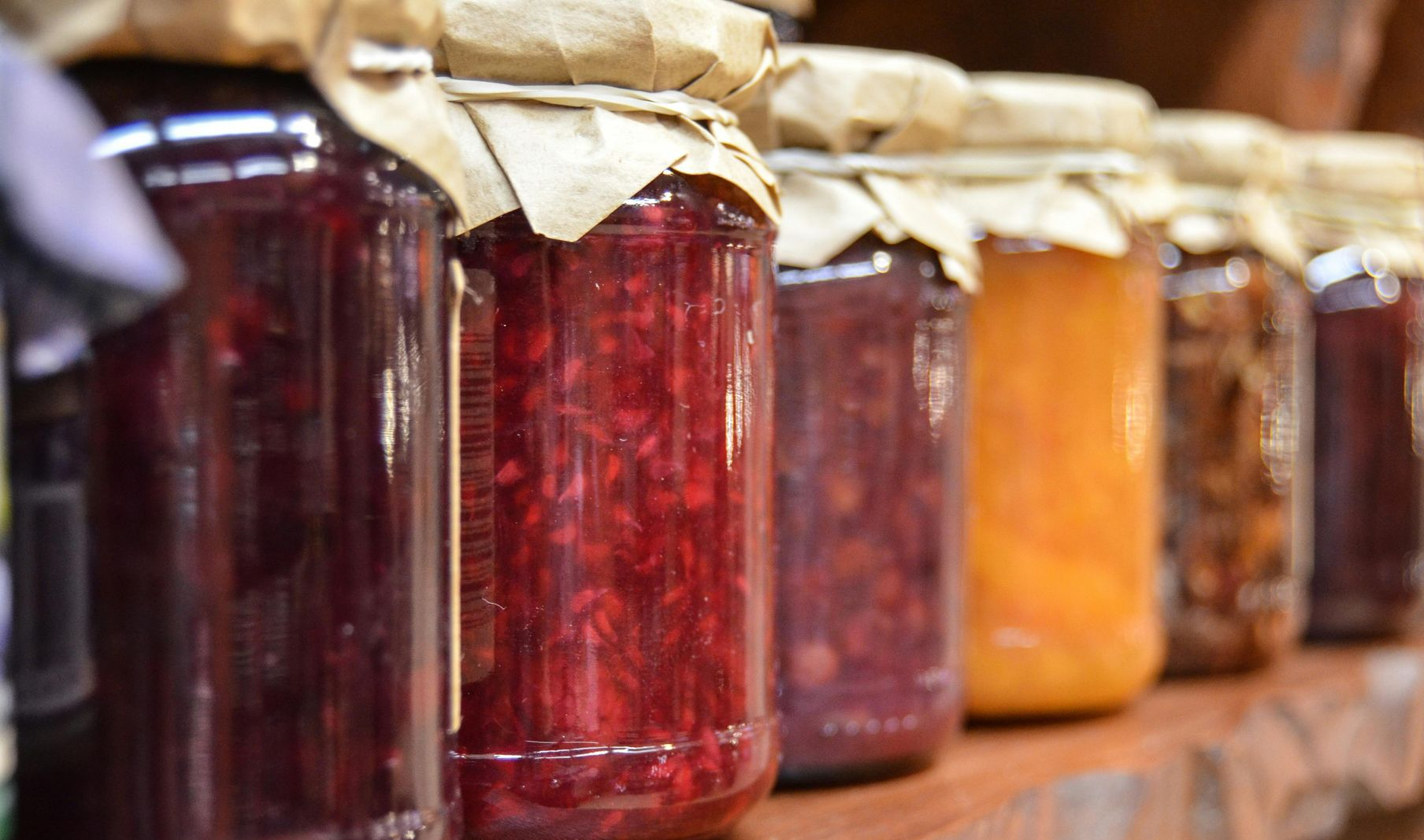 Row of glass jars filled with various colorful preserves, sealed with paper covers, on a wooden shelf.