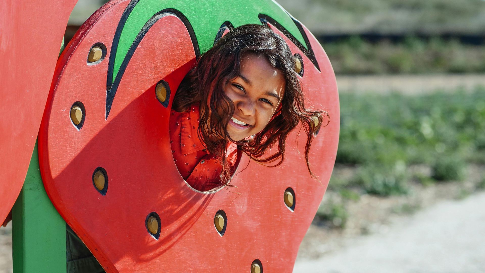 Girl peeking through a red strawberry cutout, smiling, outdoors.