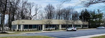 Single-story beige office building with large windows beside a road and parked car, under leafless trees