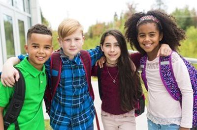 Four children standing side-by-side with arms around each other, smiling outdoors while wearing backpacks.