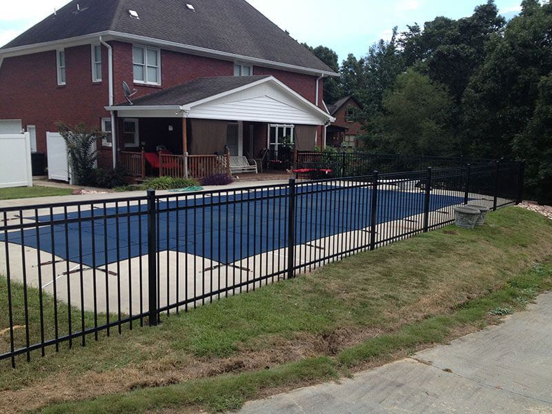 A fence surrounds a large swimming pool in front of a brick house