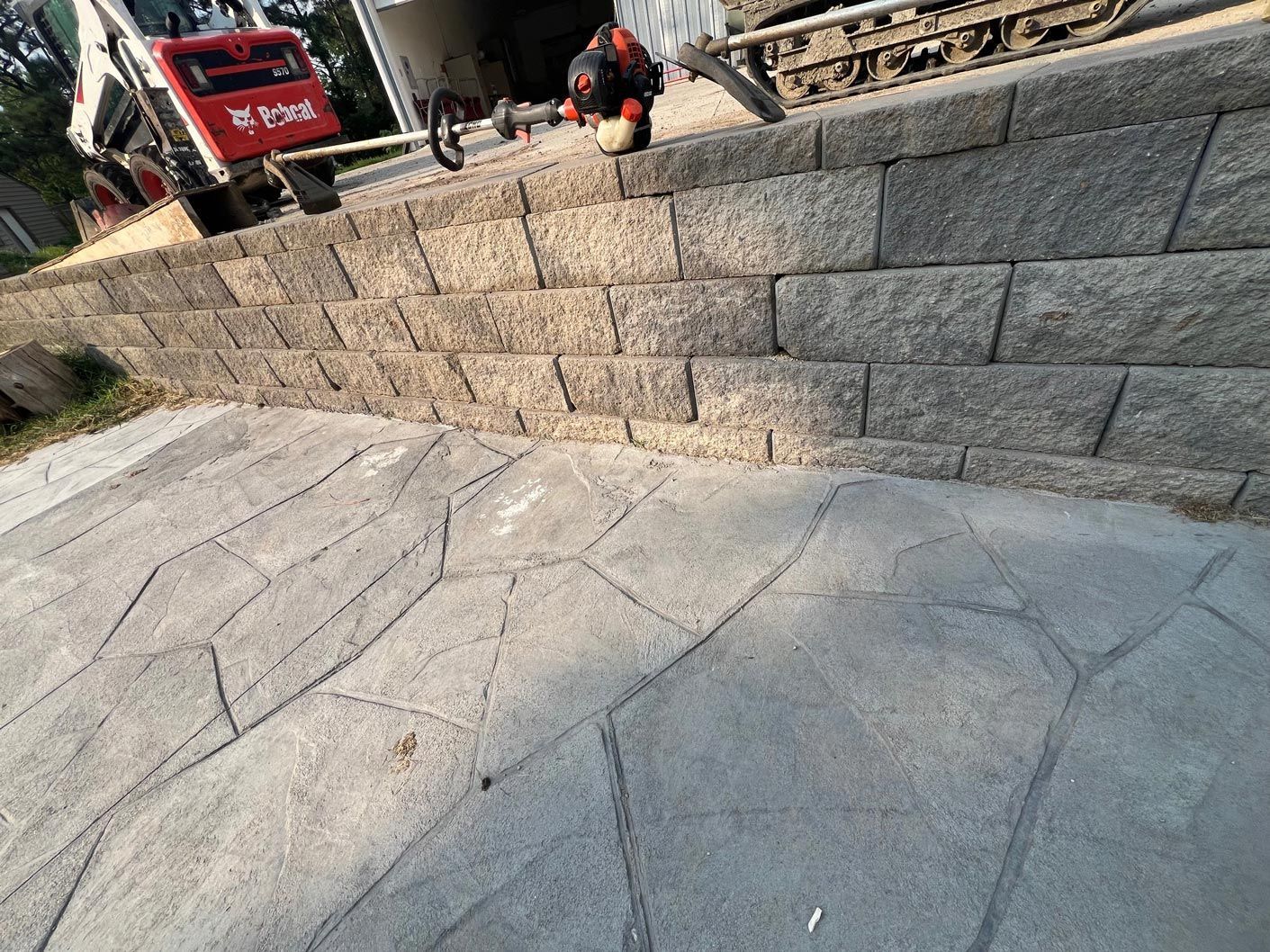 Stone retaining wall with a stamped concrete surface below; machinery visible in background.