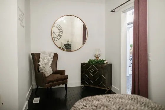 A cozy room with a brown armchair, decorative cabinet, large round mirror, and dark flooring.