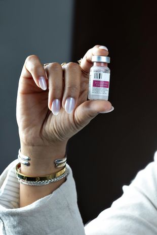 Hand holding a vaccine vial, with jewelry and light-colored nails.