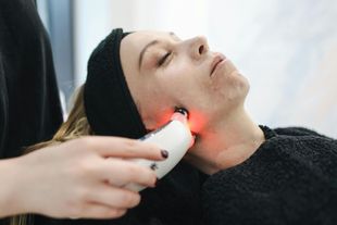 Woman receiving facial treatment with a red light device; indoor setting.