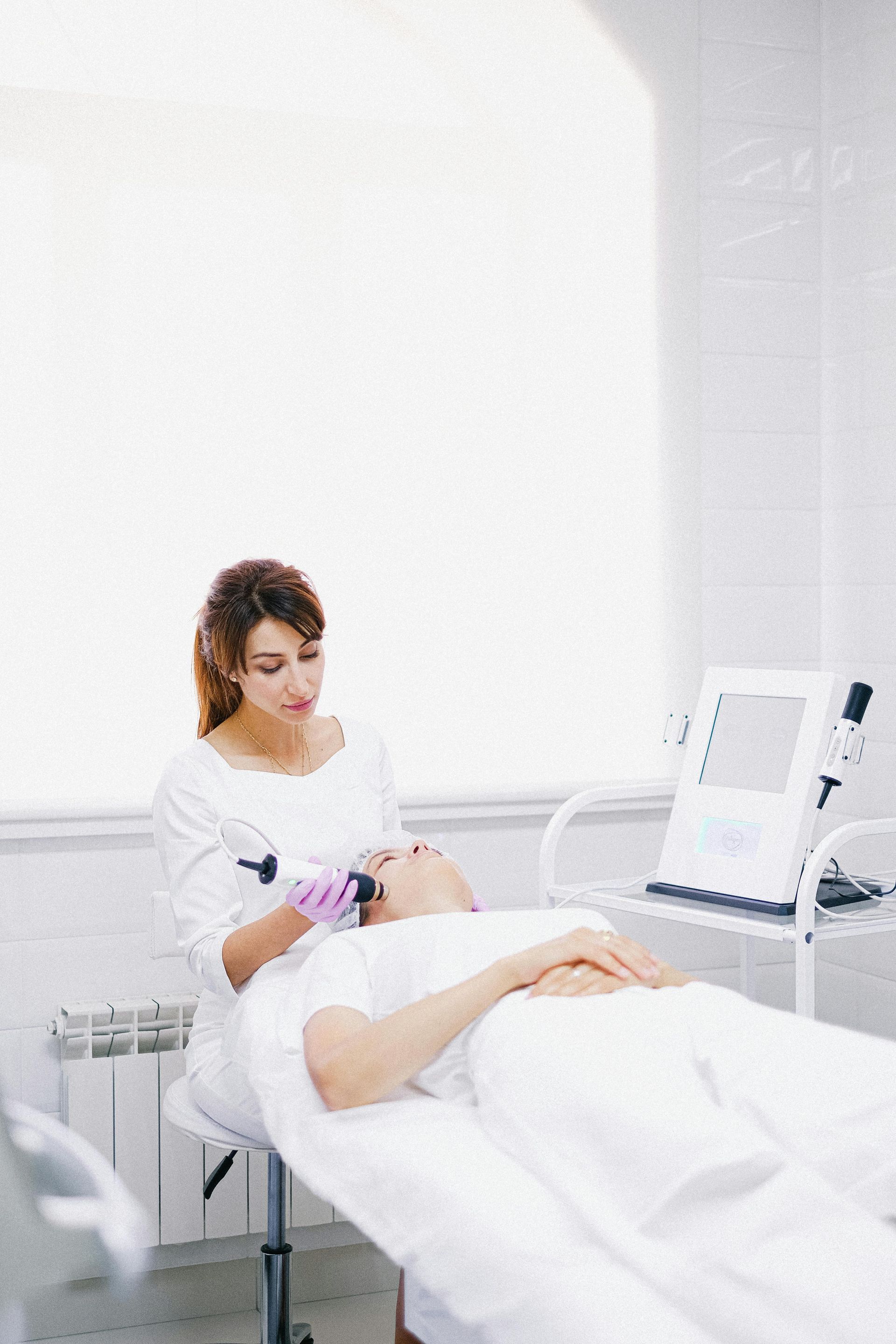 Woman performing facial treatment on a person lying on a white bed in a bright room.
