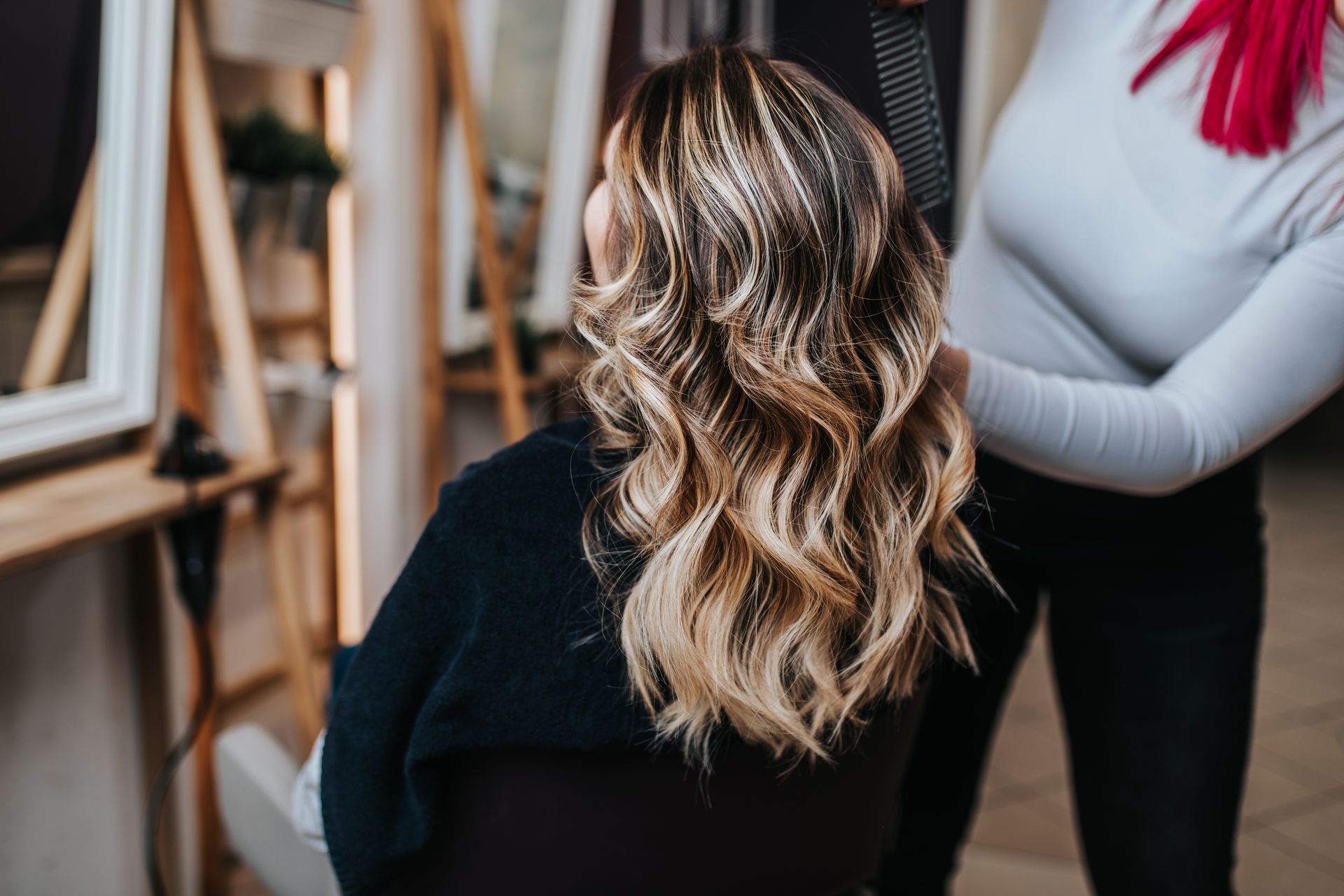 Woman with wavy blonde hair at a salon. Stylist touches hair, mirrors and tools in the background.