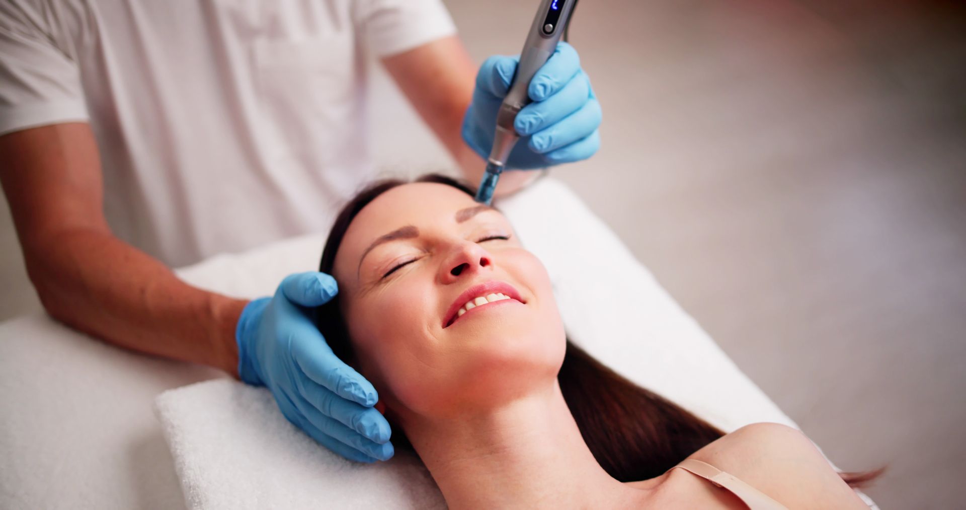 Woman receiving facial treatment; therapist using handheld device. Blue gloves, white setting.