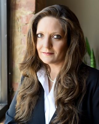 A woman in a black jacket and white shirt is standing in front of a window.