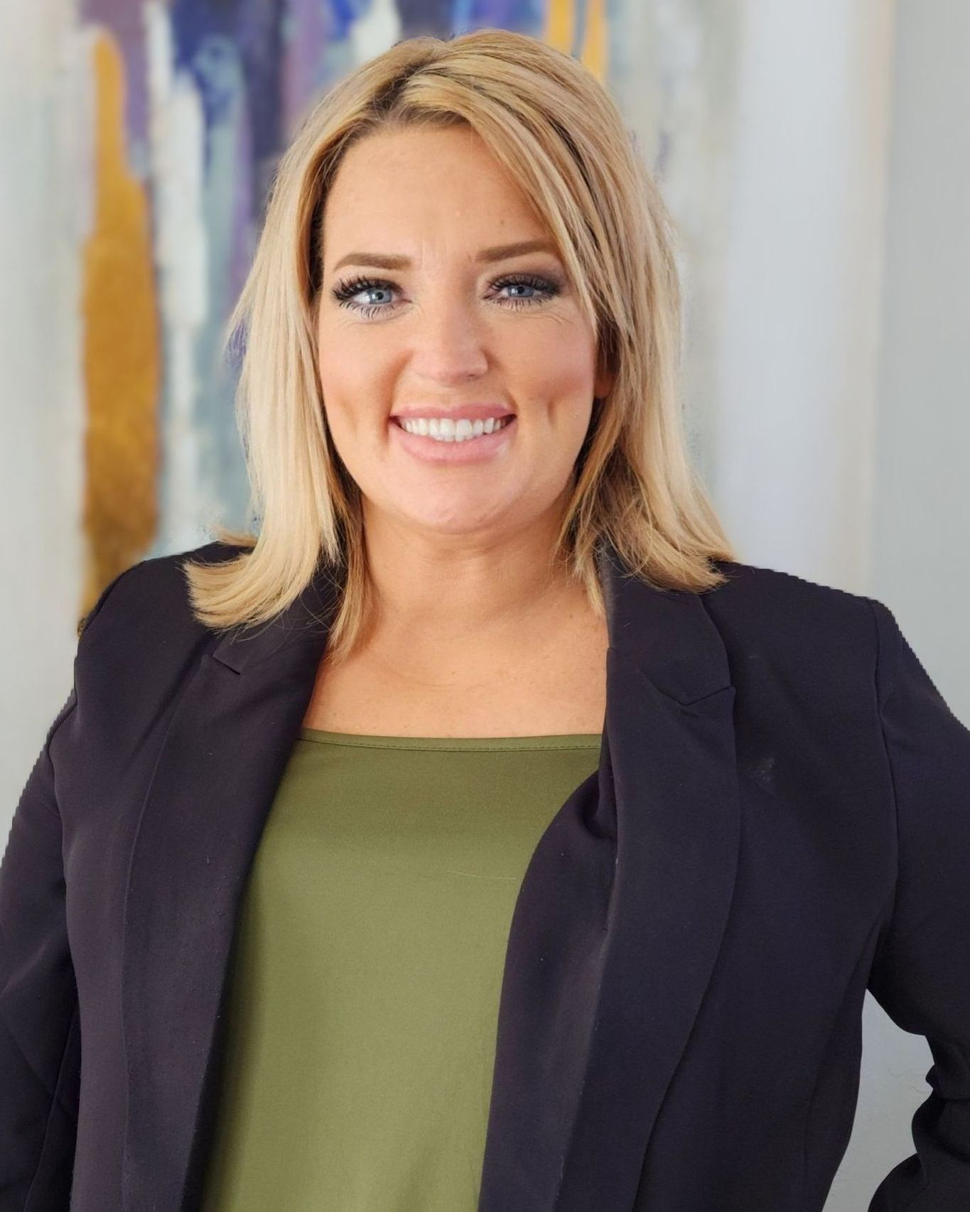 A woman wearing a black jacket and a green shirt is smiling in front of a painting.