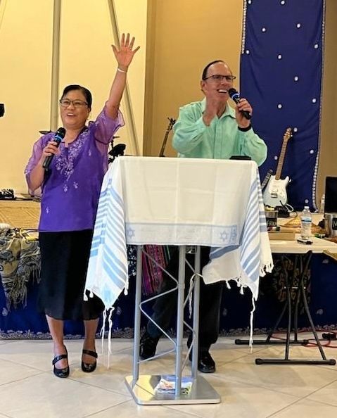 Two people stand at a podium with microphones, one raising a hand, both smiling. Blue and white cloth is draped on a table.