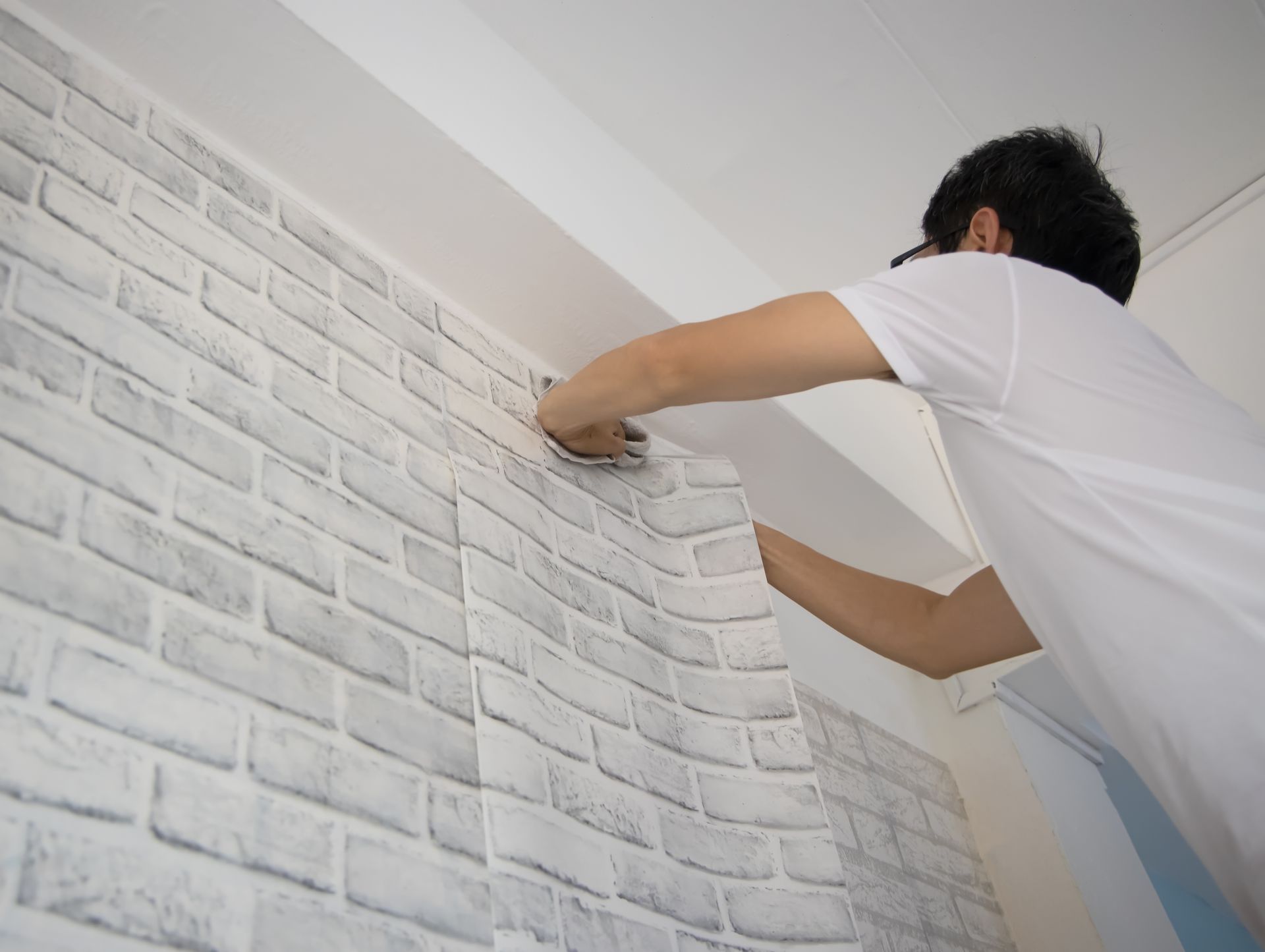 A man is applying wallpaper to a white brick wall.