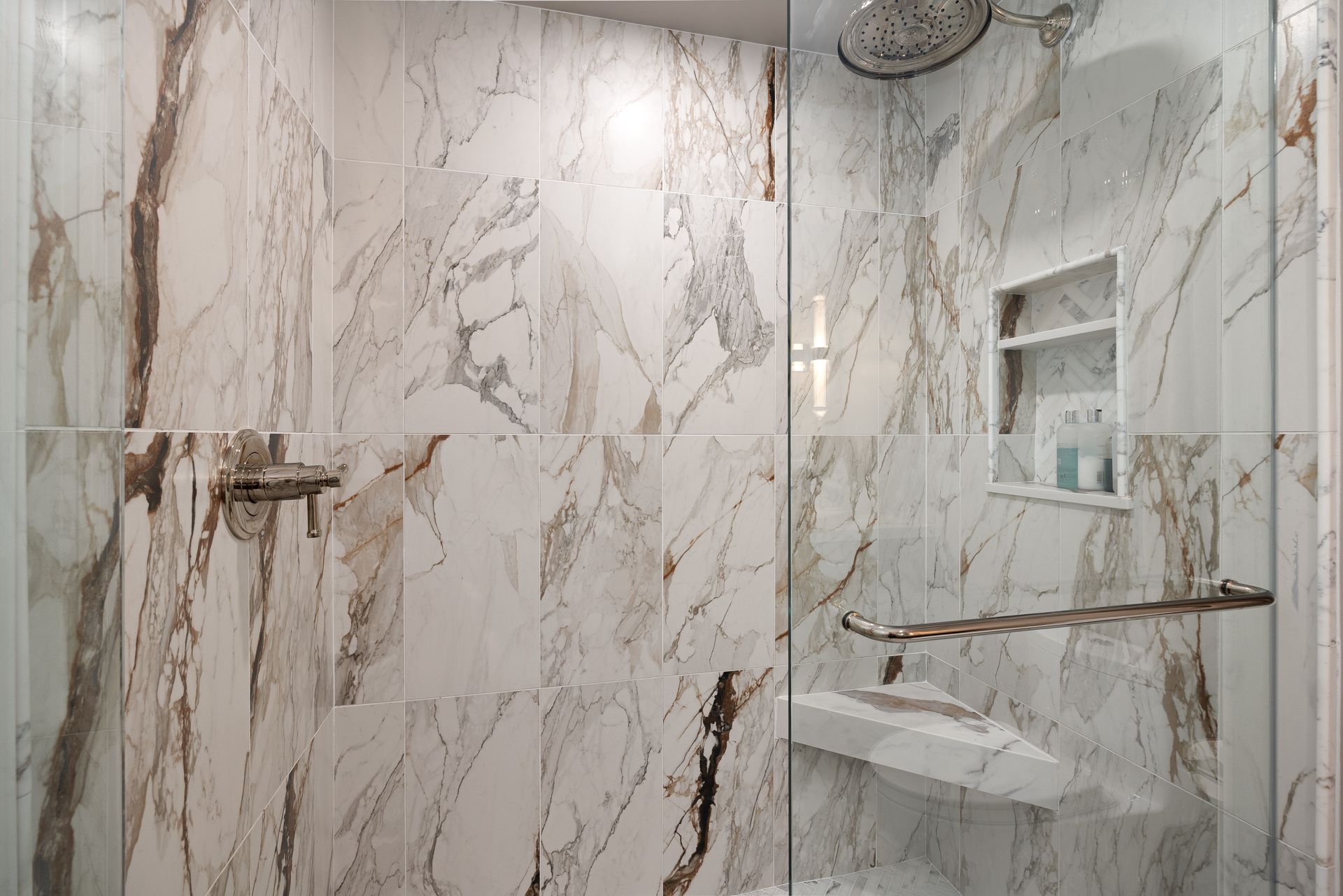 Marble-tiled shower with glass door. Includes a rain shower head and built-in shelf with toiletries.