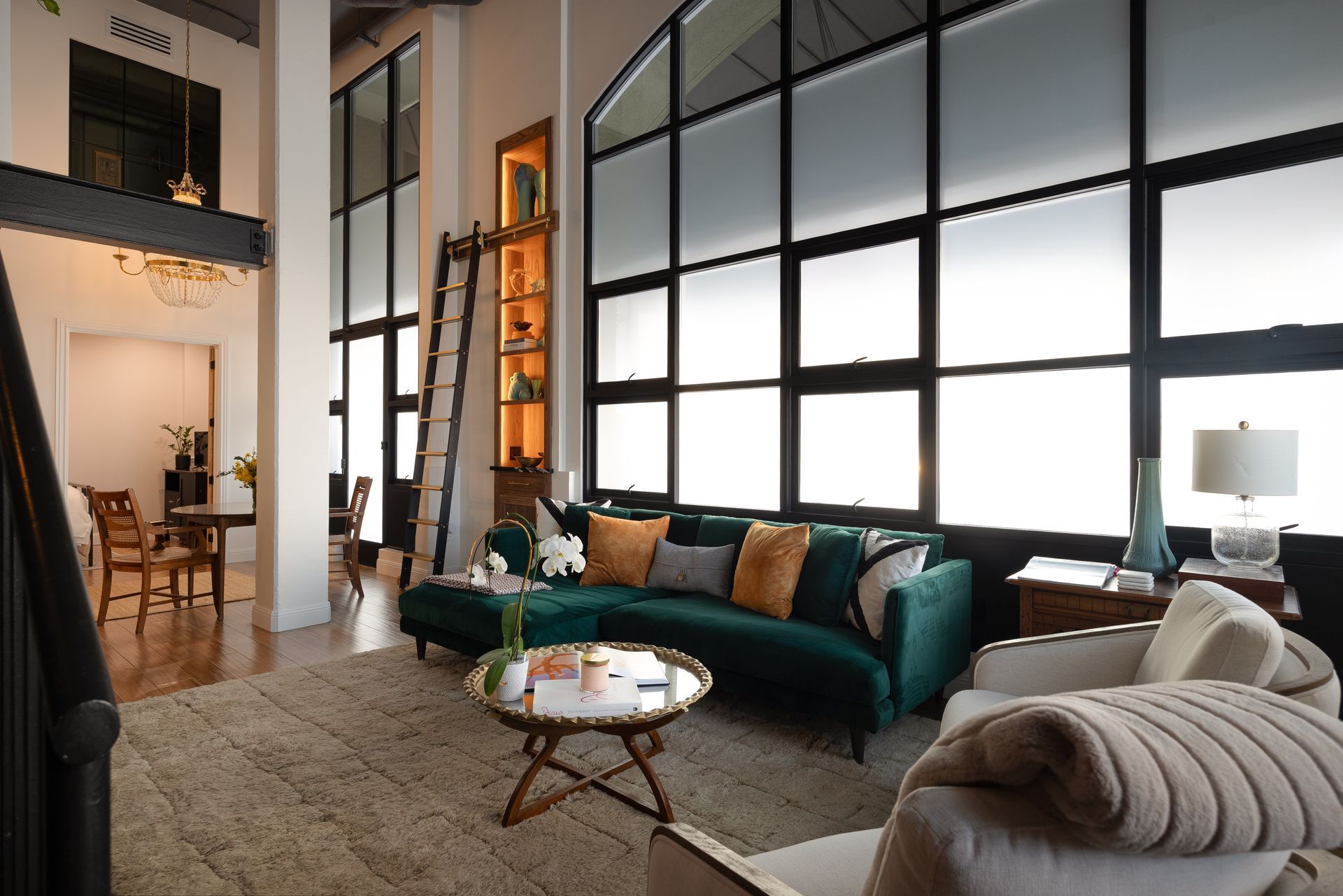 Living room with a green velvet sofa, white chairs, large windows, and a rug.