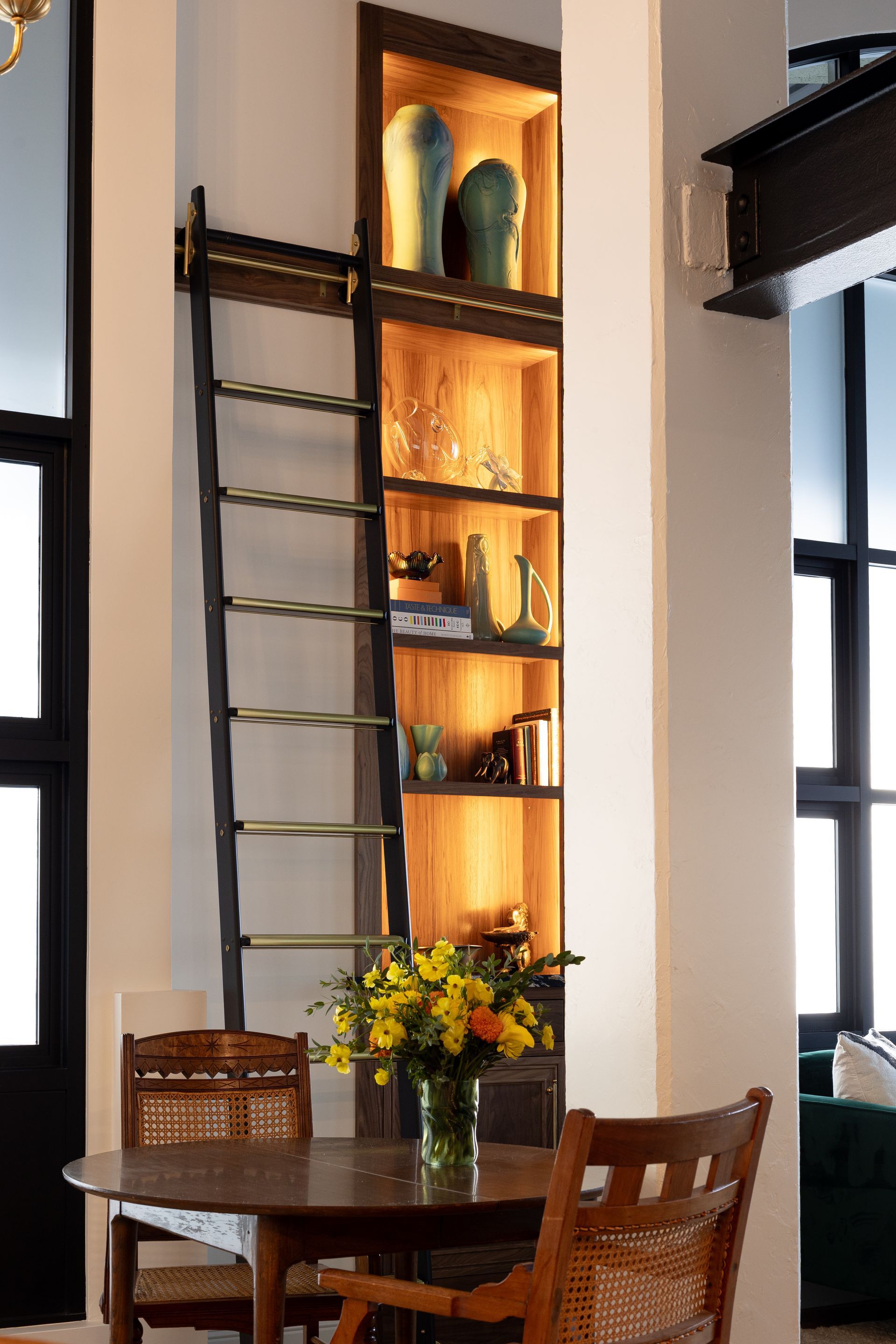 Wooden bookshelf with ladder, built into a wall. Warm lighting illuminates the shelves, displaying decor.