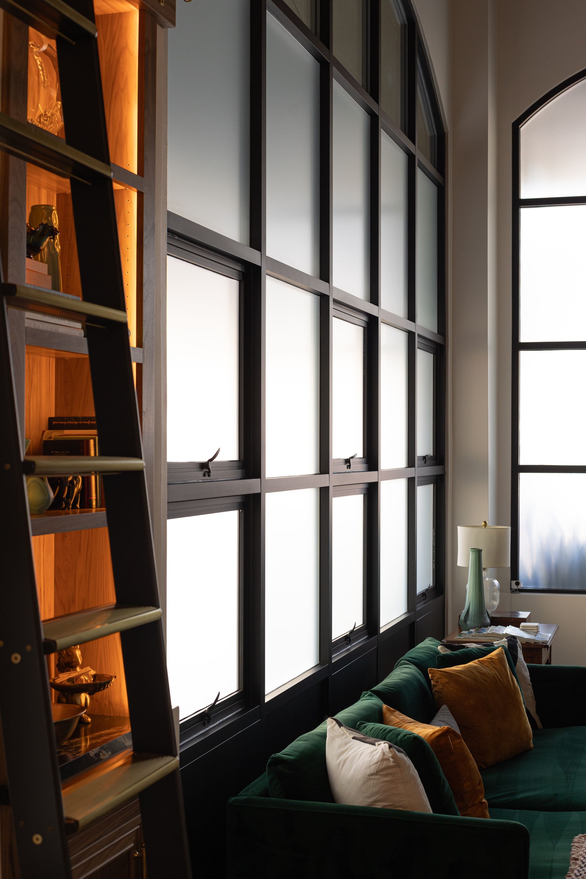 Library with dark wooden shelves, ladder, and a green velvet sofa with colorful pillows against large, frosted windows.