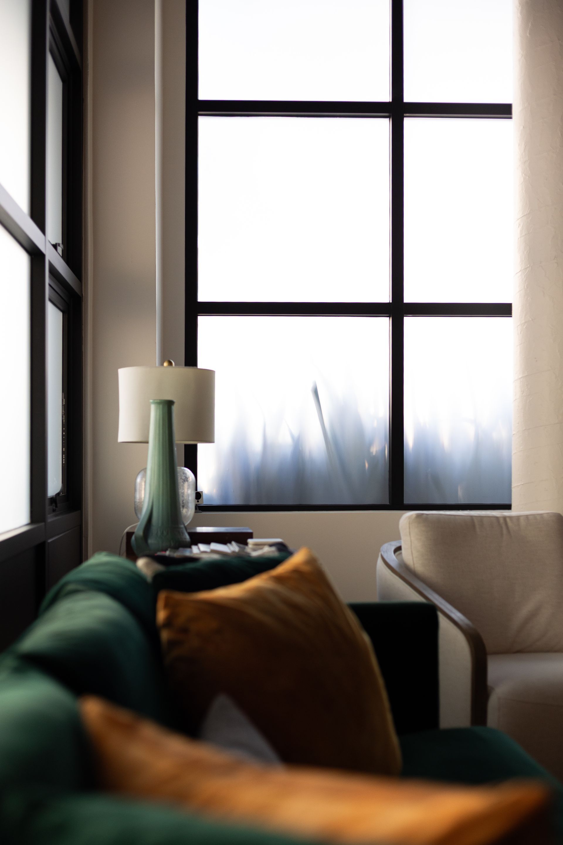 Green velvet couch, gold pillow, table lamp, and window with obscured view in a modern living room.