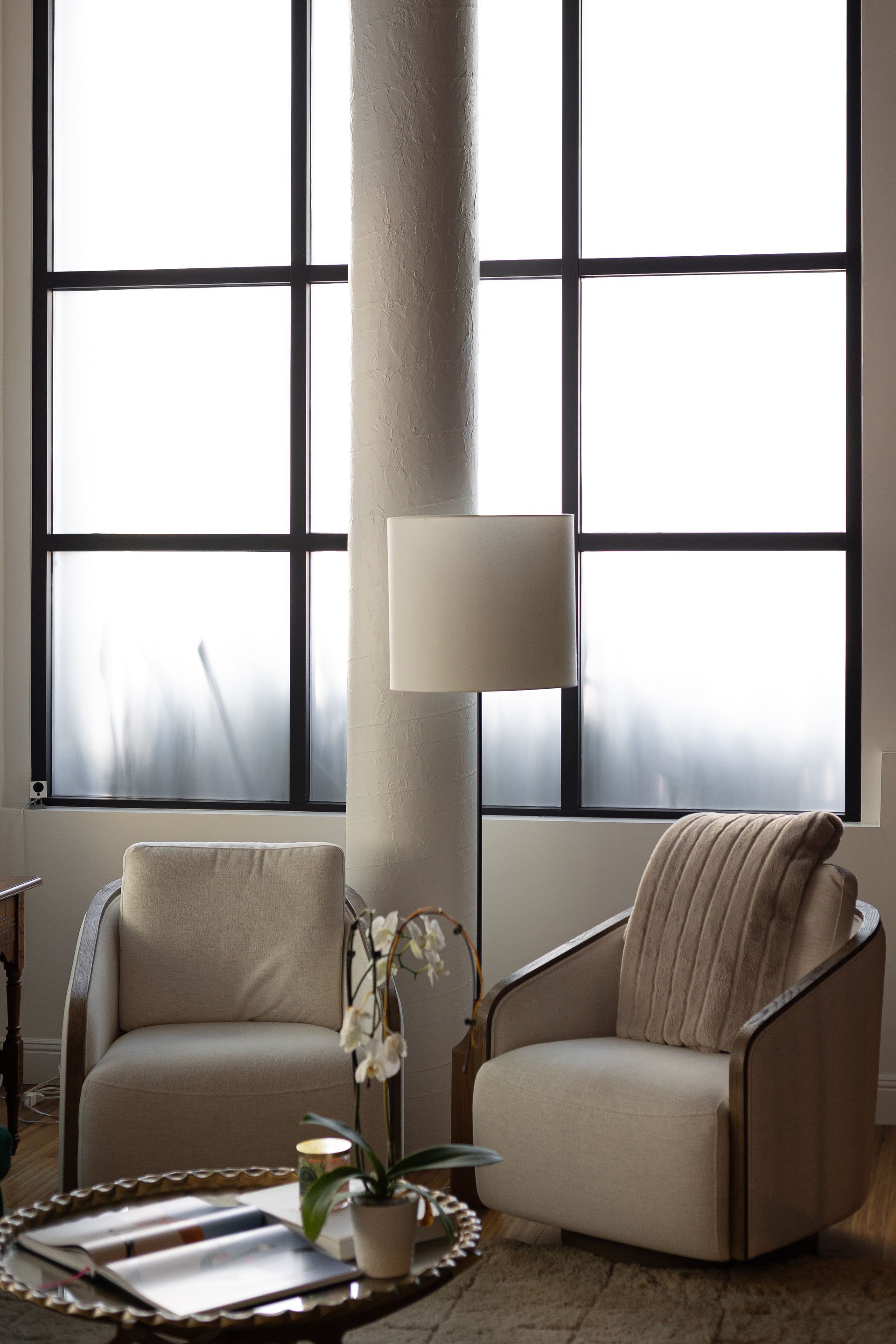 Two cream armchairs, a lamp, and a coffee table in front of a large window.