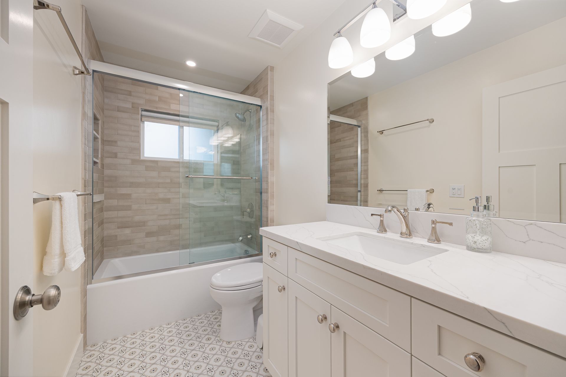 Modern bathroom with white vanity, dual sinks, glass shower, and patterned floor.