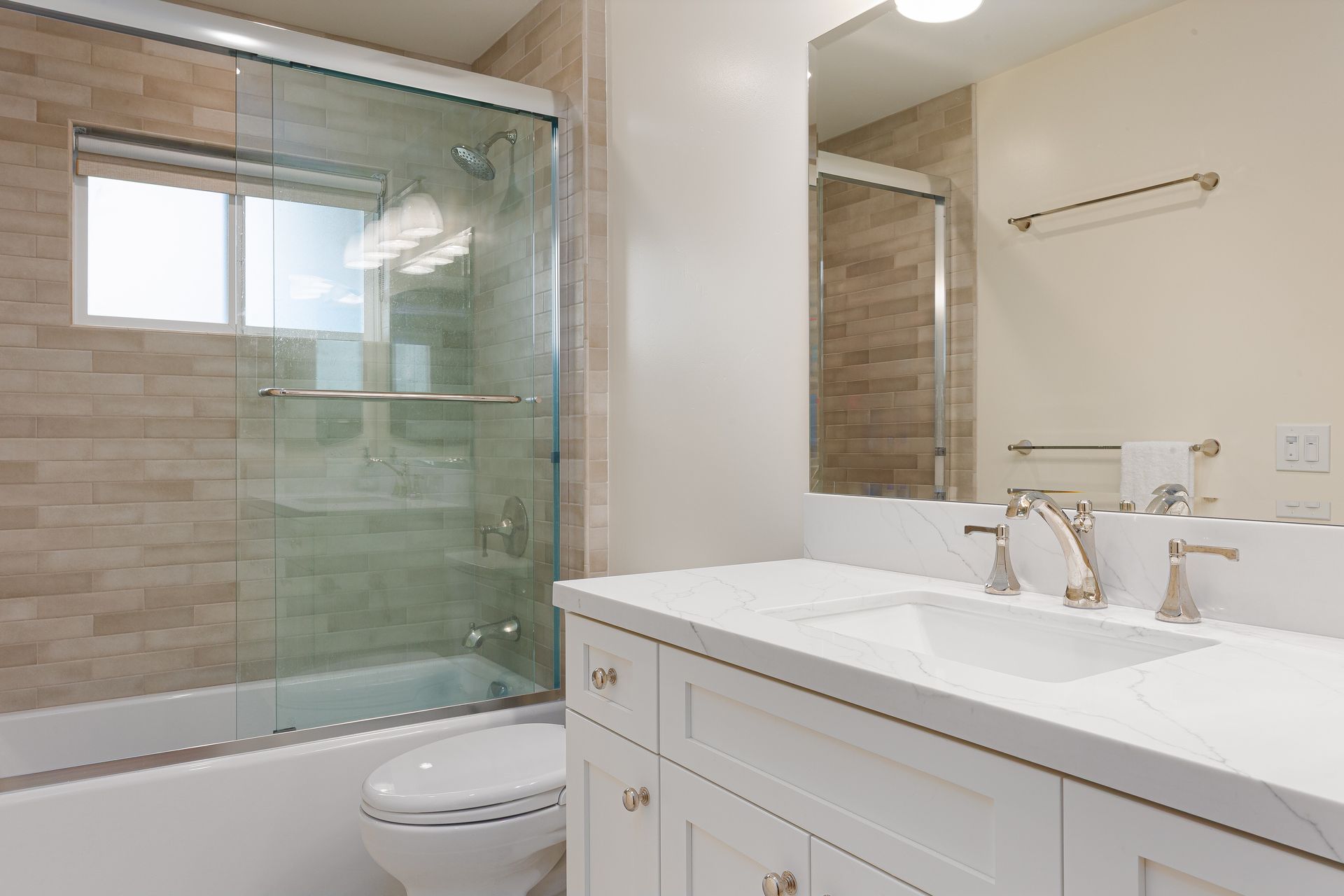 Bathroom with white vanity, toilet, and bathtub. Beige tiled walls, glass shower door, and a window.