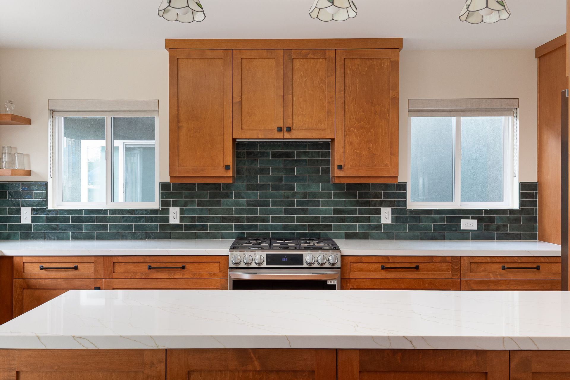 Kitchen with light wood cabinets, green tile backsplash, white countertops, and stainless steel range.