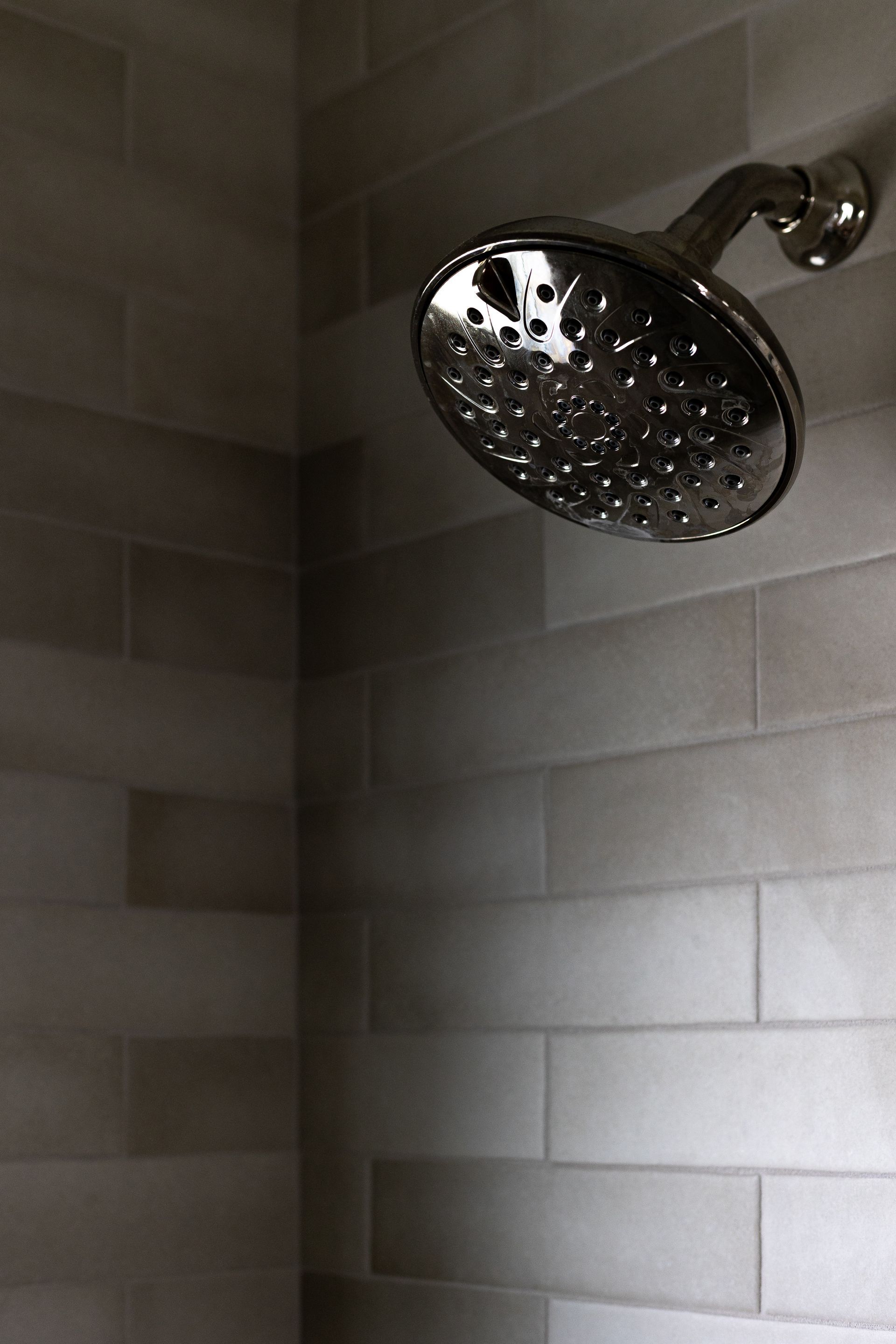 Shower head with water droplets against gray, rectangular tiled wall.