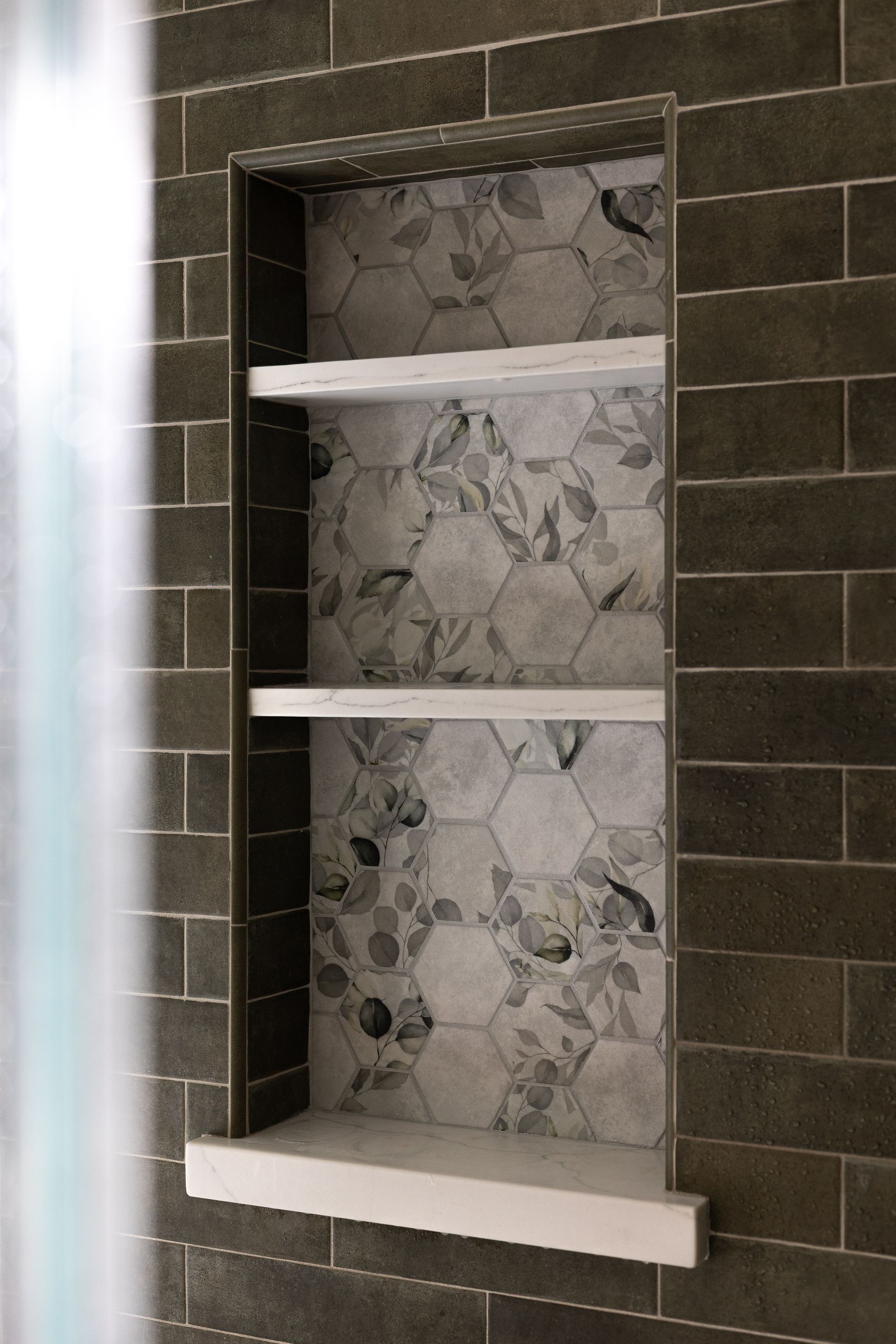 Recessed shower shelf with three white shelves. Backed by floral hexagonal tile, surrounded by dark brown rectangular tiles.