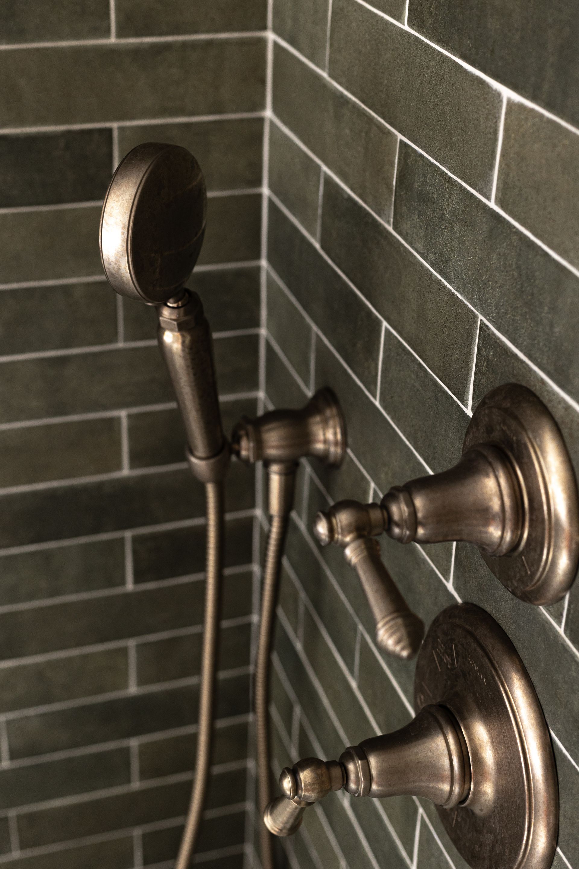 Bronze shower fixtures on dark green tiled wall. Handheld shower head and temperature controls.