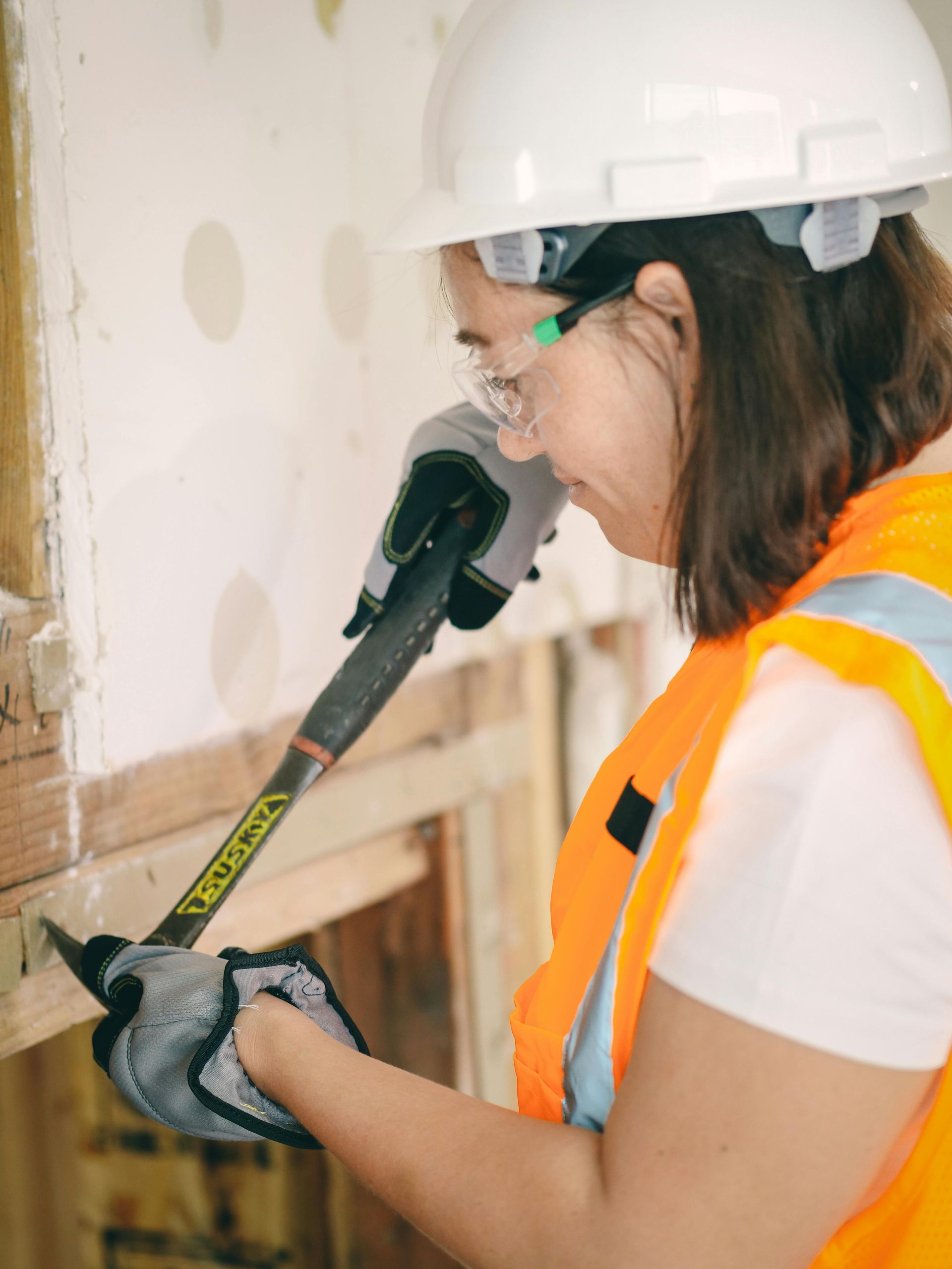 Woman in hard hat, safety glasses, and vest using a pry bar to remove wood in a construction setting.