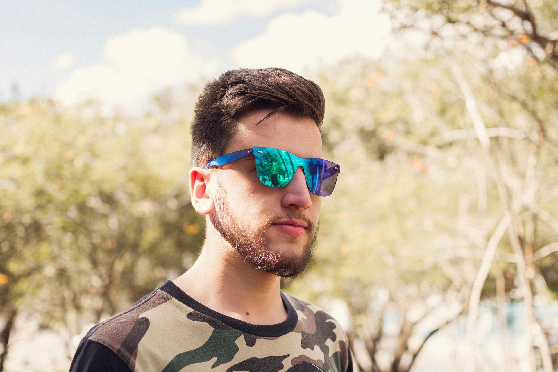 Man wearing sunglasses with reflective blue lenses and camouflage shirt outdoors.
