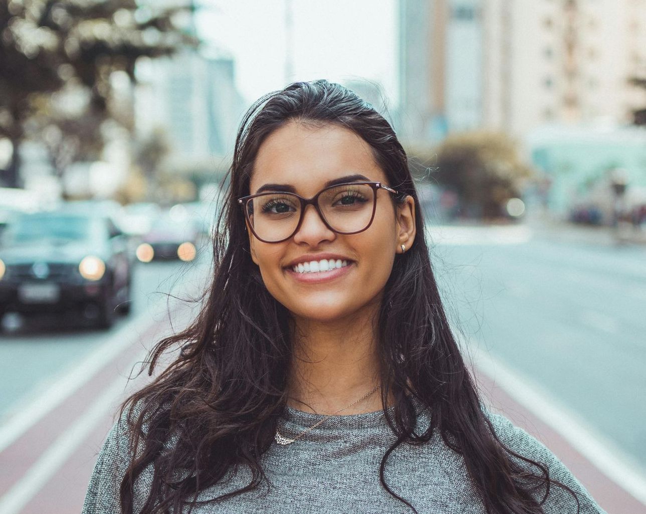 Woman with glasses smiles at the camera, standing on a city street with cars in the background.