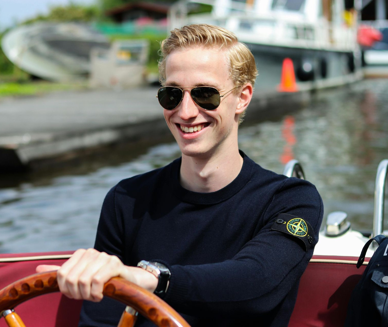 Blond man in sunglasses smiles while steering a boat. He wears a dark sweater with a logo on the sleeve.