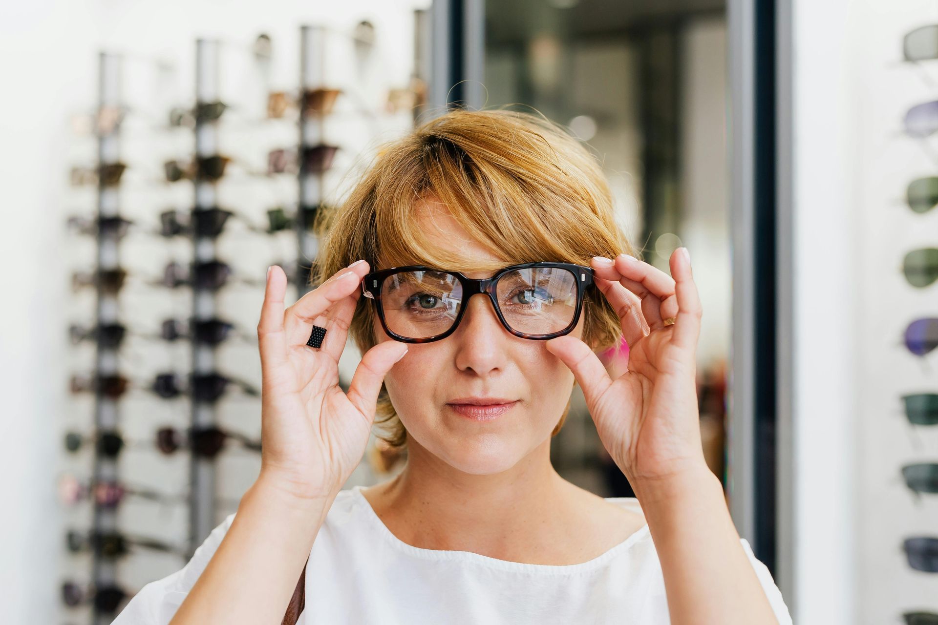 Woman in glasses looking at the camera in an eyewear store, holding the glasses with both hands.