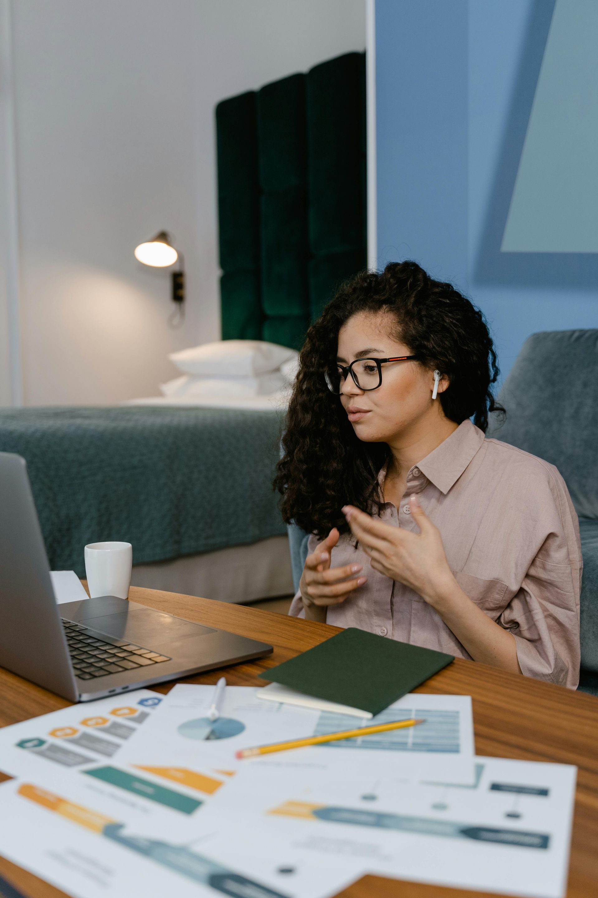 Woman with glasses in a virtual meeting, gesturing at a laptop with documents on a desk in a bedroom.