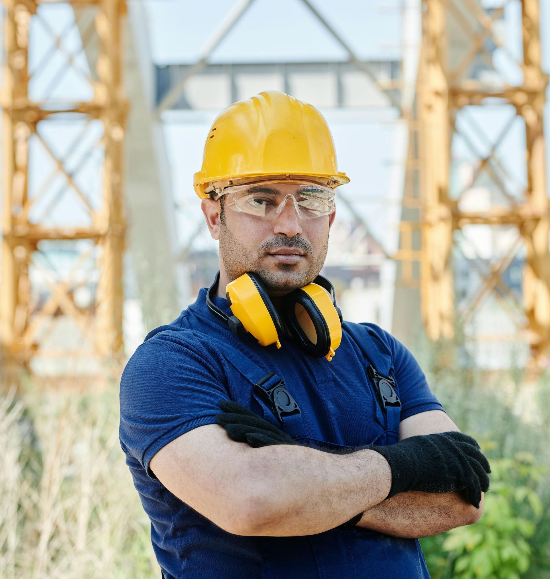 Construction worker with yellow hard hat, safety glasses, and ear protection, arms crossed on site.