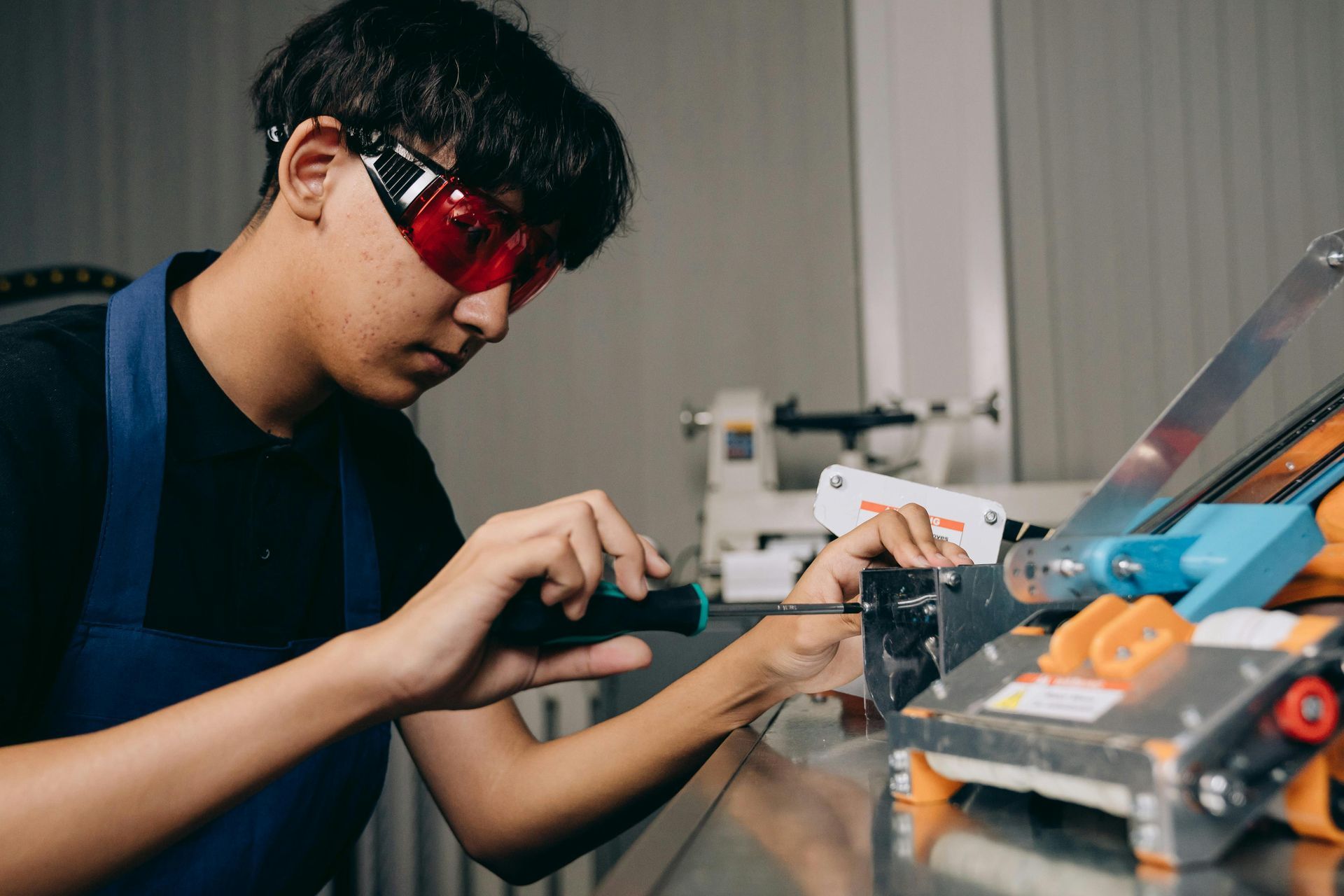 A young man with safety glasses uses a screwdriver on a metal machine in a workshop.