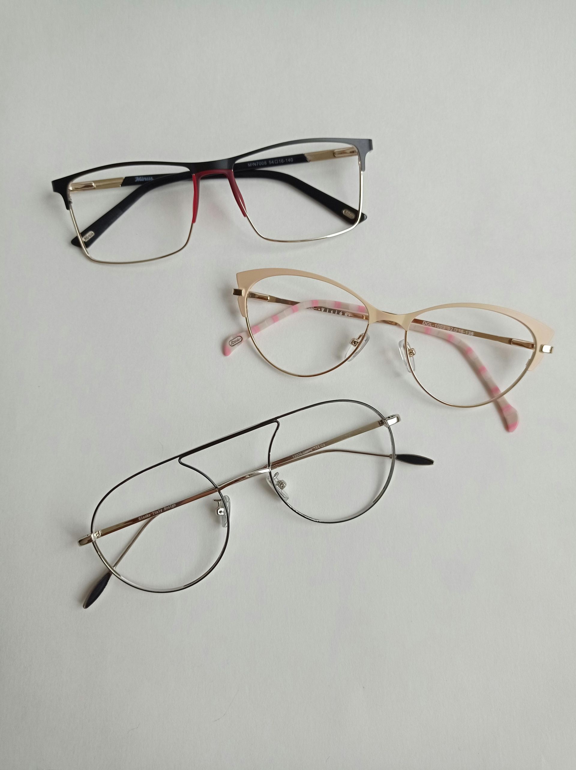 Three pairs of eyeglasses on a white surface: black and gold rectangle, cat-eye, and round with a double bar.