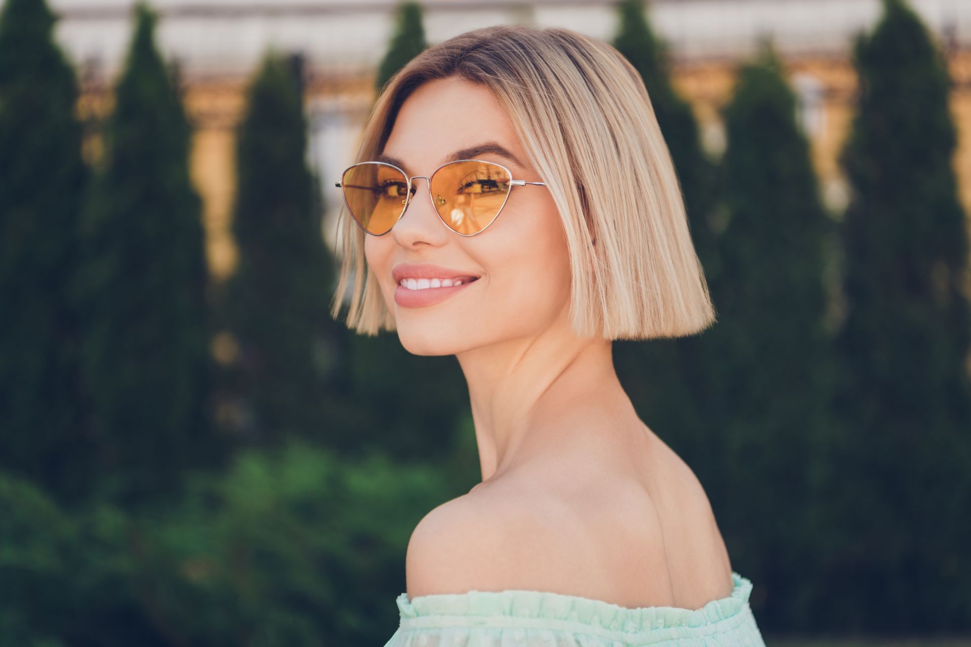 Woman with blonde bob and orange sunglasses smiles, looking over her shoulder outdoors.