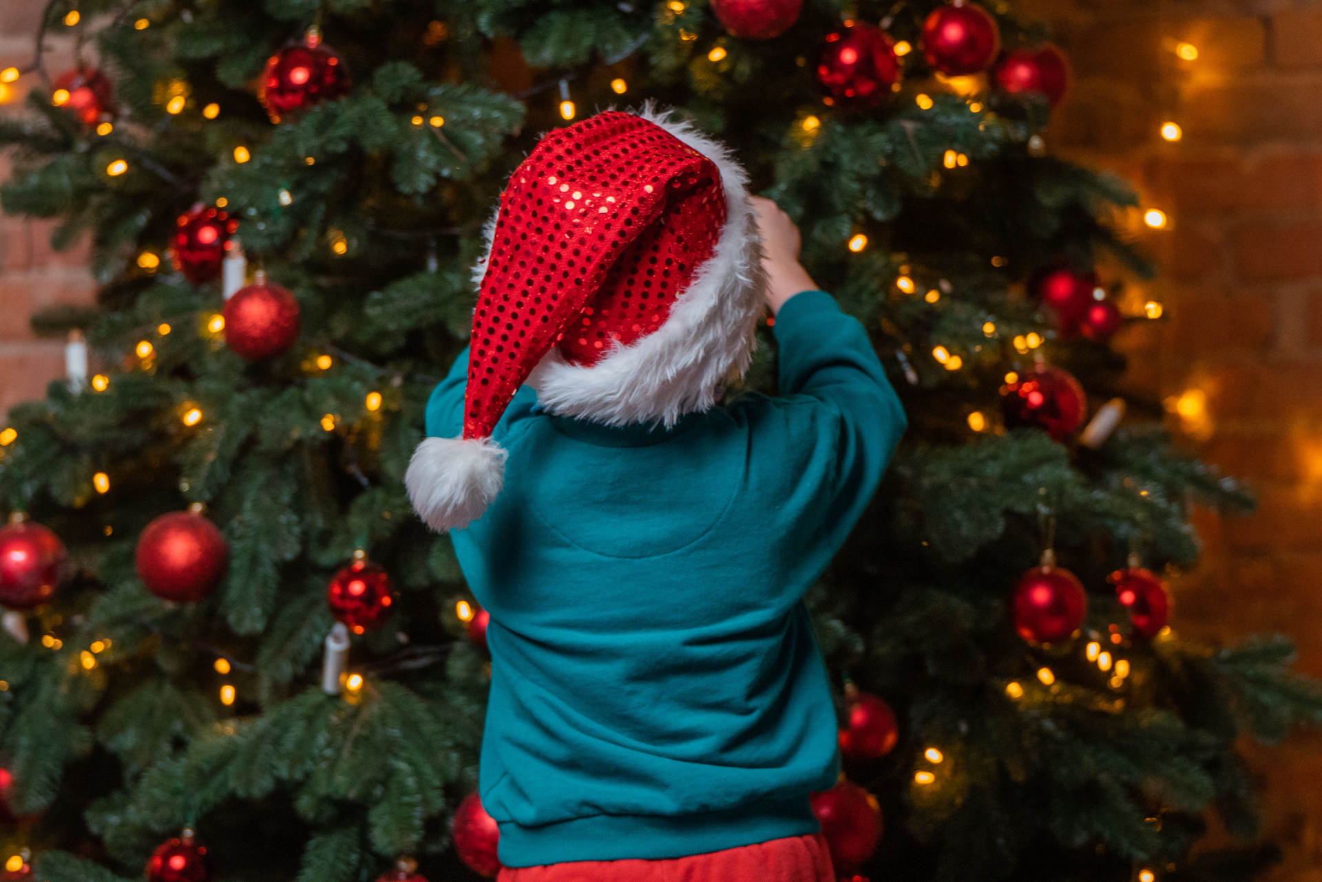 Criança usando chapéu de Papai Noel em frente a uma árvore de Natal com enfeites e luzes.